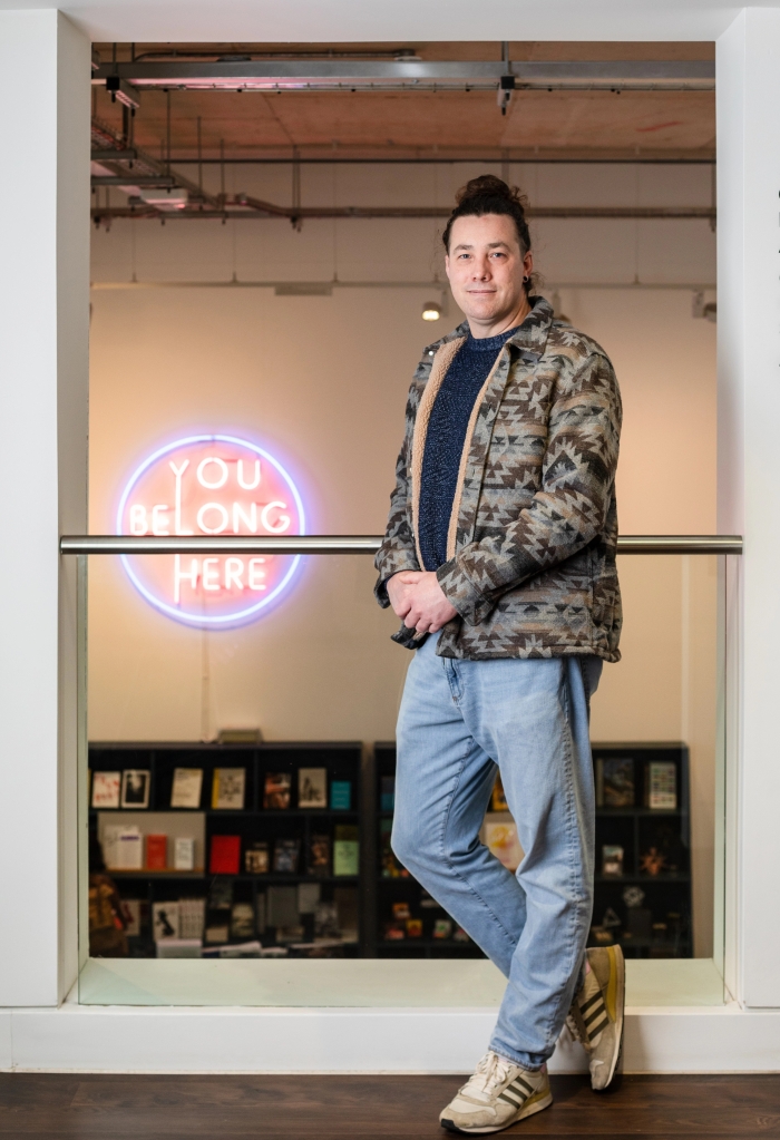 A full length portrait of Liam, a white male with brown hair tied back in a bun. He is dressed in a patterened grey and brown fleece; a blue jumper; light blue jeans; white trainers. In the background is foyer of John Hansard Gallery, Southampton and an artwork made of neon lights with the text "You Belong Here"